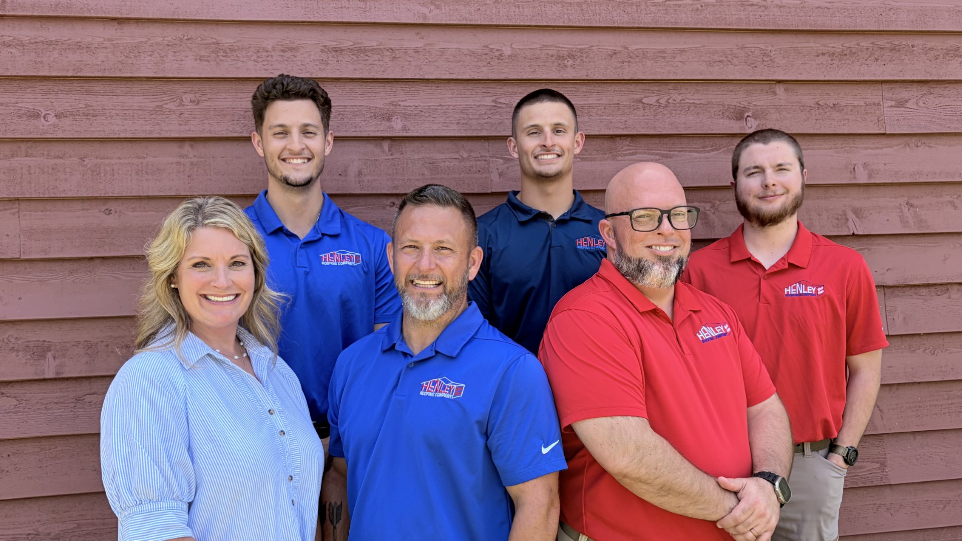 Six adults, four men and two women, pose and smile in front of a wooden wall; the men wear red and blue shirts with logos.