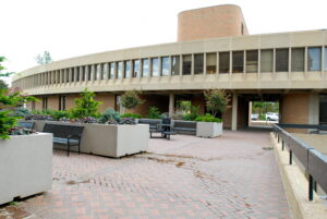 A modern building with large windows and a brick facade overlooks a courtyard with benches, planters, brick paving, and some greenery.