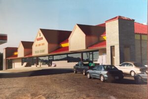 A row of cars is parked outside a shopping center with a red roof and a sign that reads 
