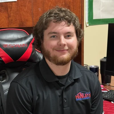 A man with curly hair and a beard, wearing a black Henley Roofing Company polo, sits in an office chair at a desk.