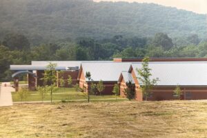 A group of brick school buildings with gray metal roofs sits in front of a wooded hillside and grassy foreground.