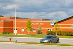 A gray car parked in front of Brickey-McCloud Elementary School, a red brick building with a green lawn and a partly cloudy sky.