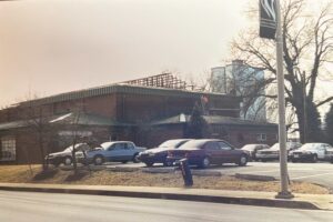 Several cars are parked in front of a brick building with a partially constructed roof; a worker is visible on the roof, and leafless trees stand nearby.