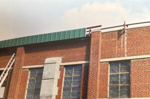 A brick building with large windows, a green metal roof edge, and a ladder leaning against the side. Some metal rails are visible on the roof.