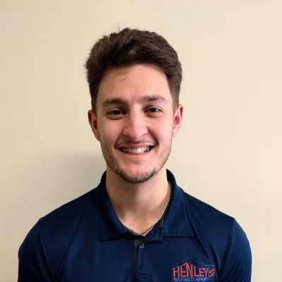 A young man with short brown hair smiles while wearing a navy blue Henley Roofing Company polo shirt, standing against a plain beige wall.