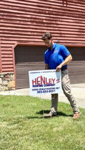 A man in a blue shirt places a Henley Roofing Company sign in the front yard of a house with a brown garage door.