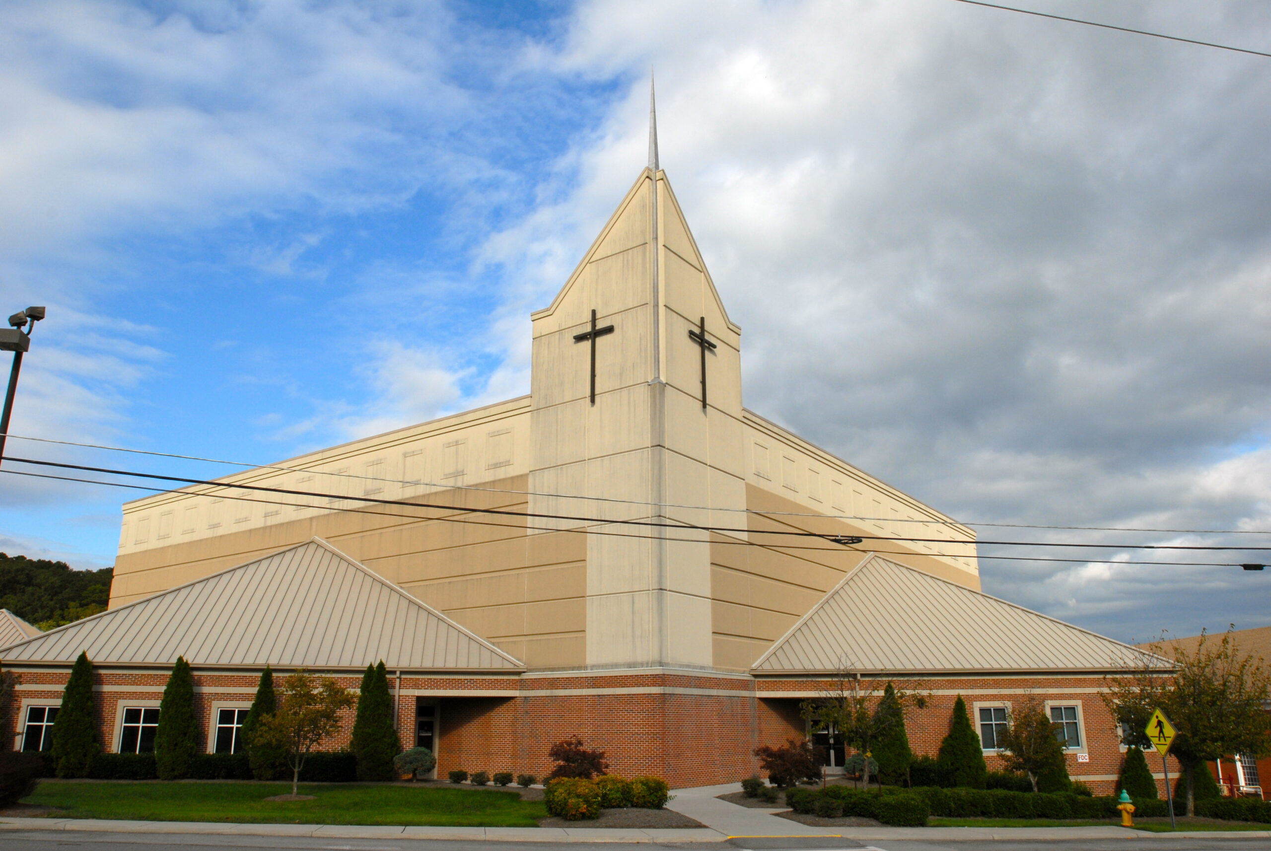 A large beige and brick church building with a tall central tower featuring a cross, set against a partly cloudy sky.