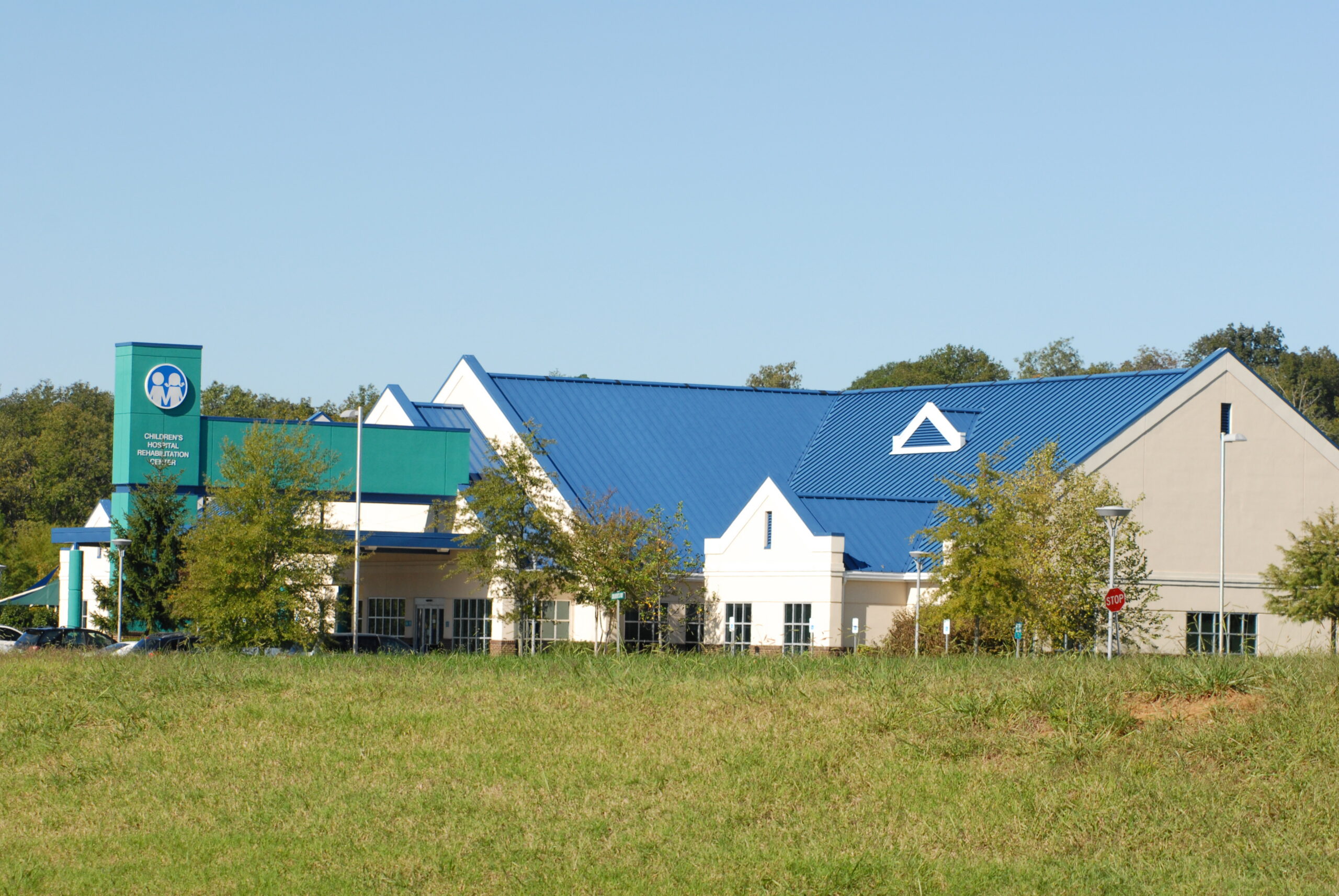A modern building with a blue roof, white walls, and a green sign, surrounded by trees and grass under a clear sky.