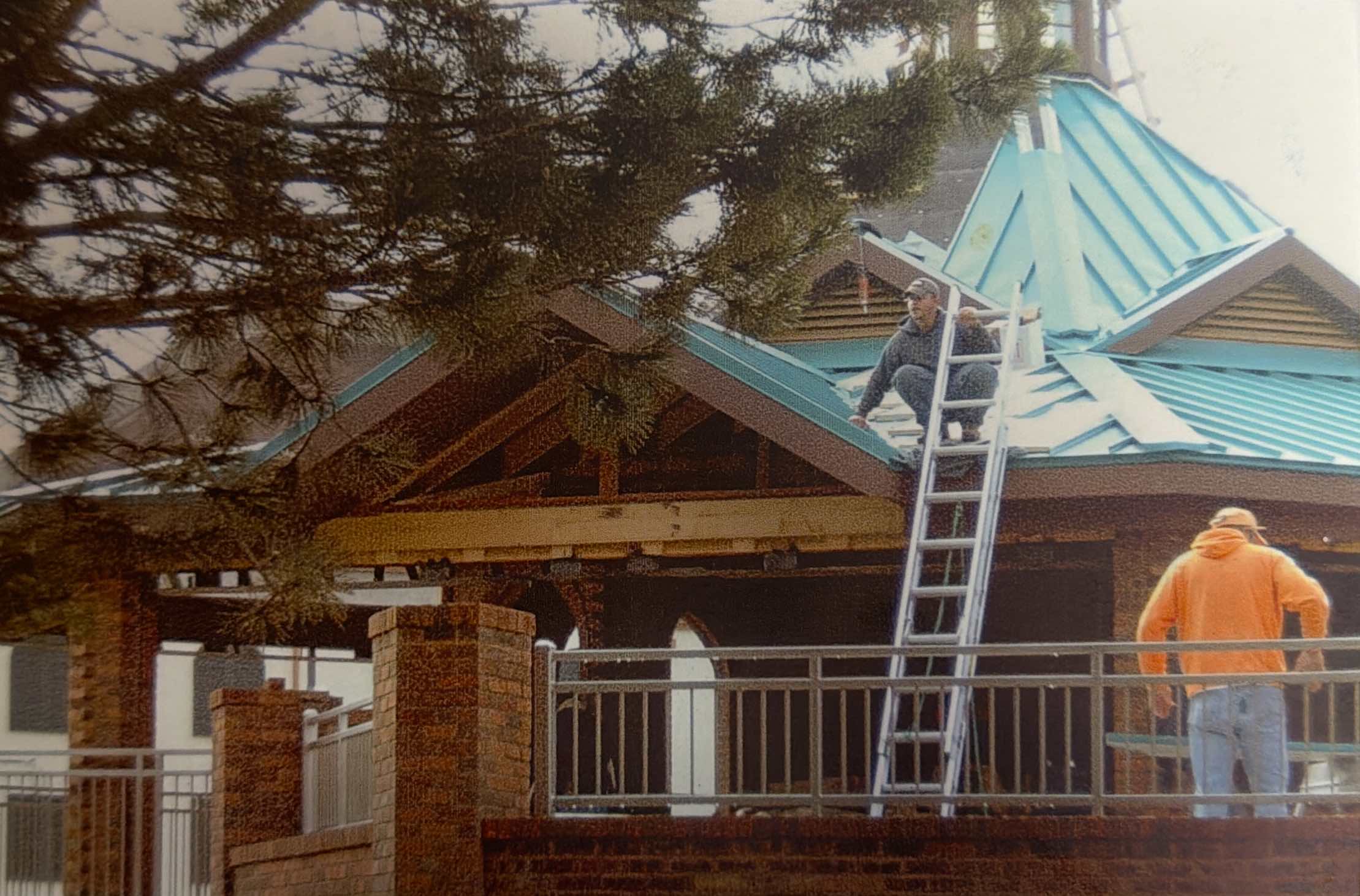 Two workers repair the roof of a brick building using a ladder, with one person on the roof and the other standing below; tree branches are visible in the foreground.