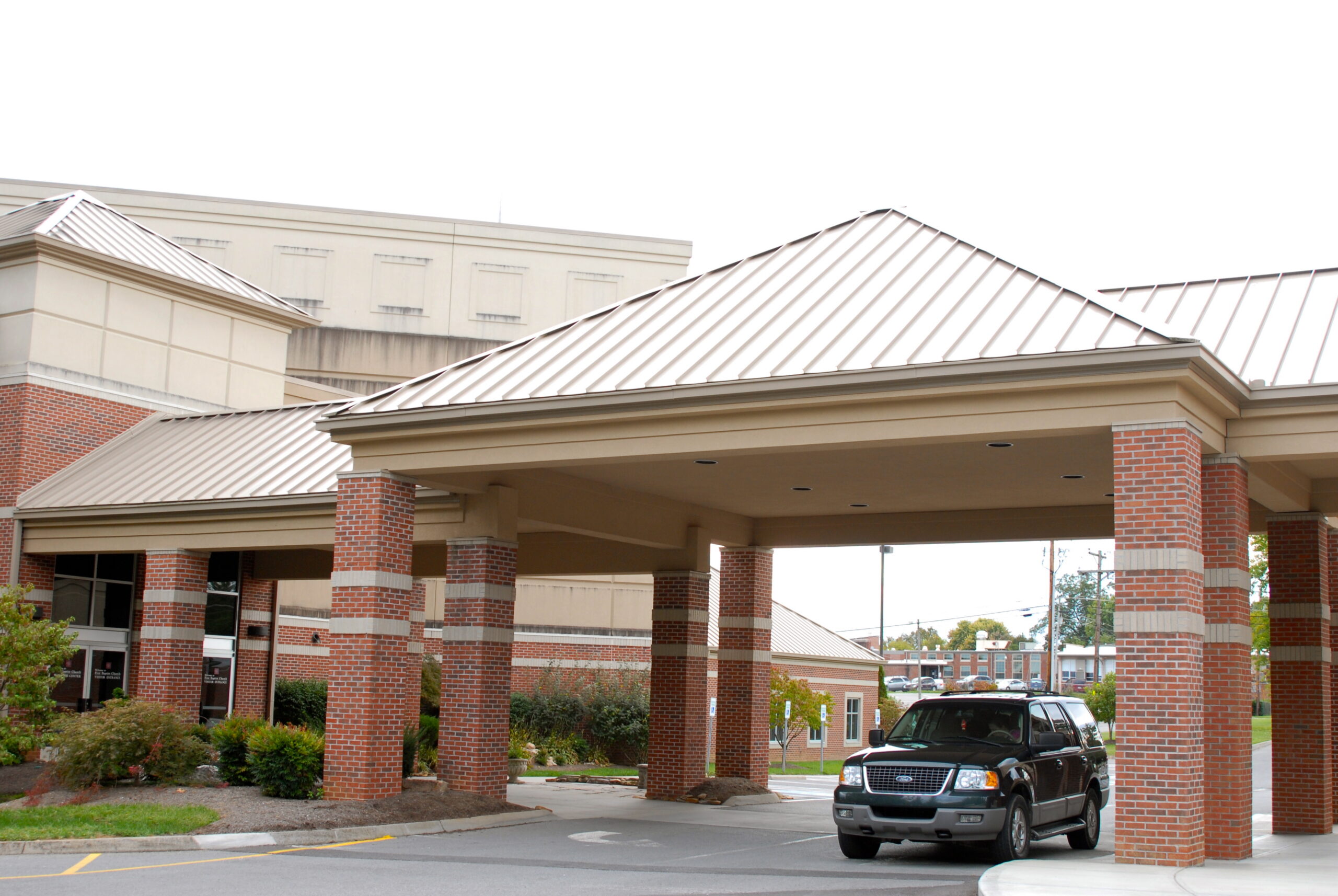 A black SUV is parked under the covered entrance of a brick building, possibly a hospital or medical facility, on a cloudy day.