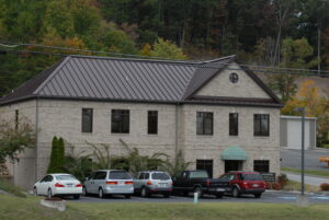 A two-story beige brick office building with several parked cars in front, surrounded by trees with autumn foliage.