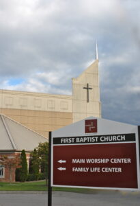 A sign in front of First Baptist Church directs to the Main Worship Center and Family Life Center; the church building and cloudy sky are visible in the background.