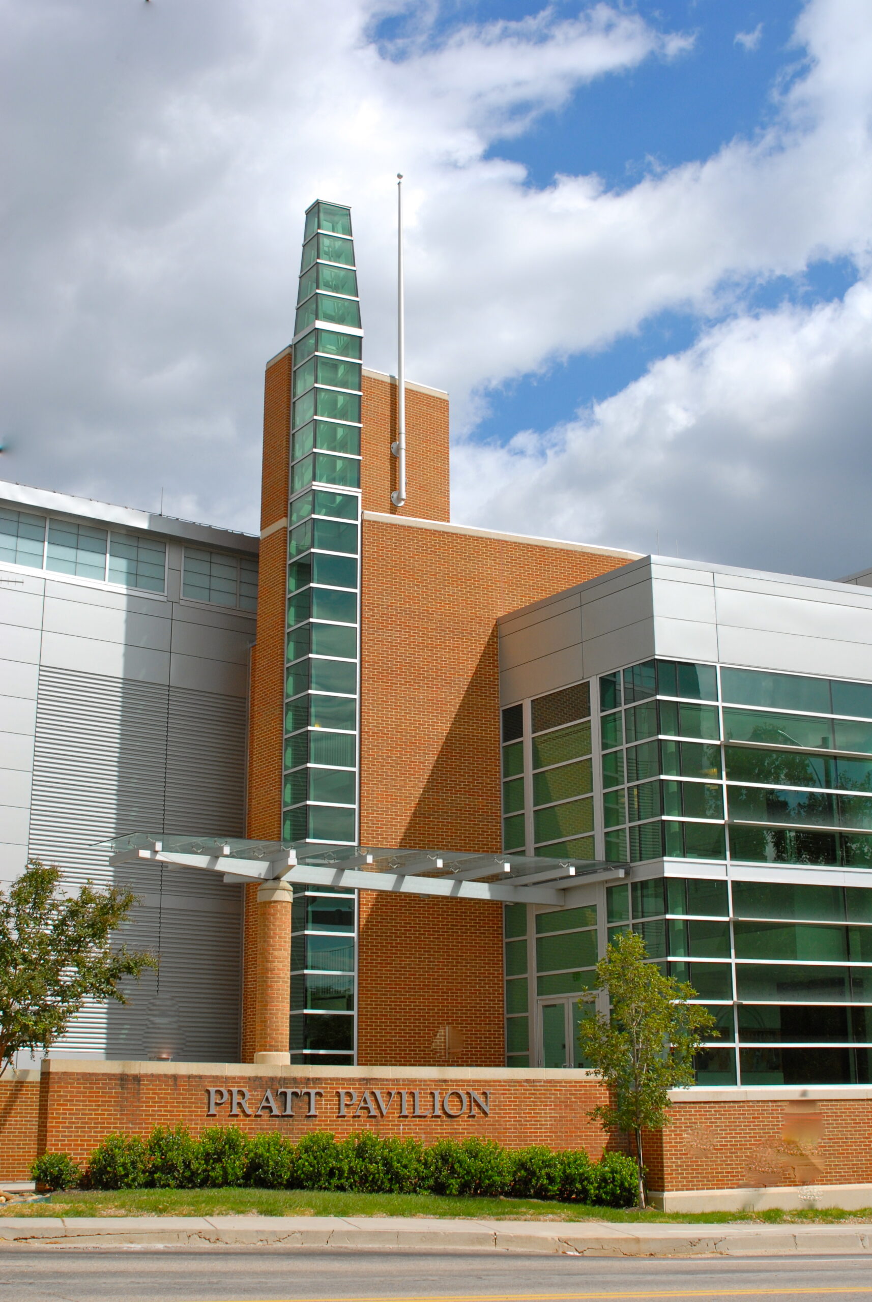 Exterior view of the Pratt Pavilion, a modern building with brick and glass architecture, featuring a tall glass tower and a sign above the entrance.
