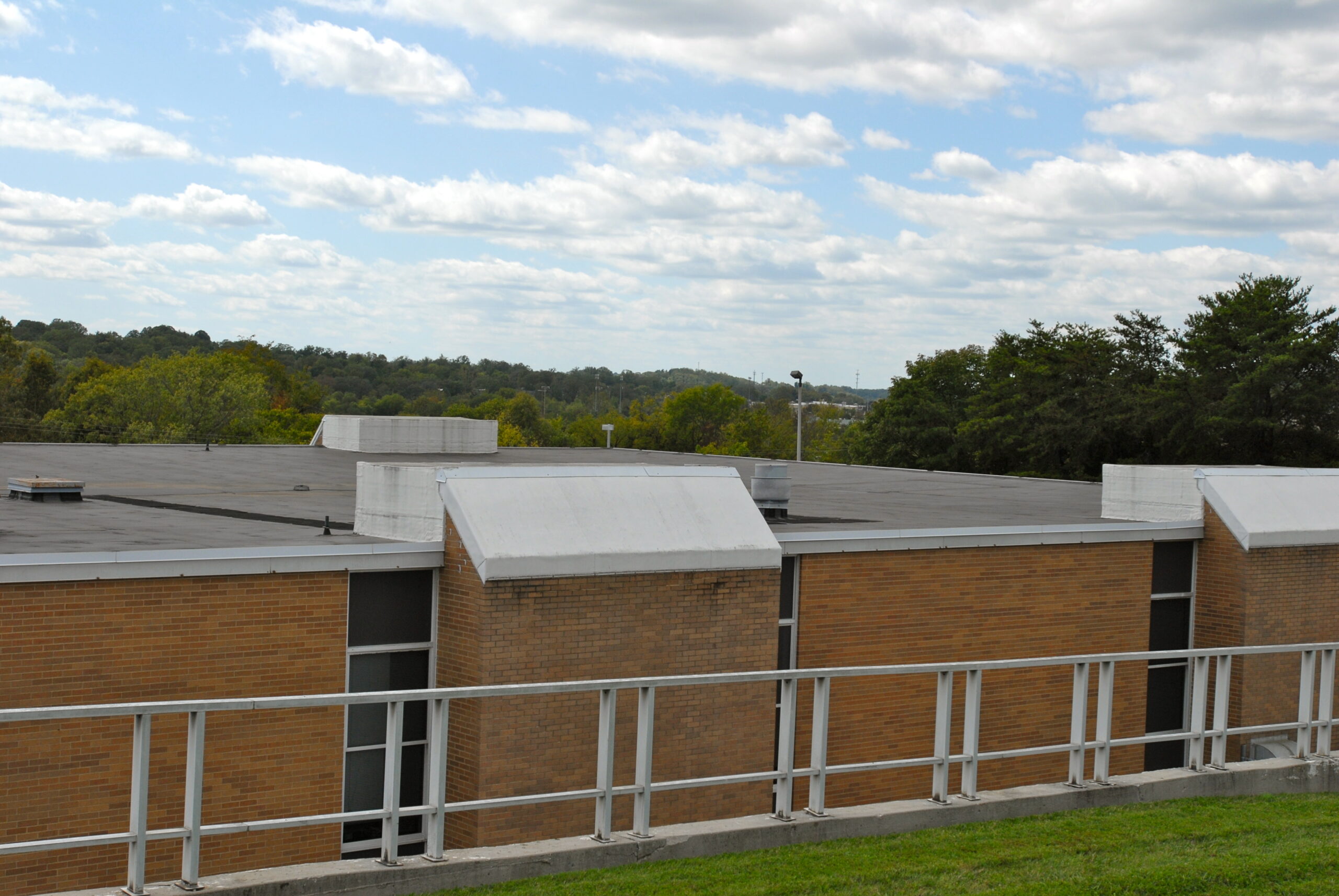 A flat-roofed brick building with white trim stands behind a metal railing, with trees and cloudy sky in the background.