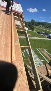 A view from a roof under construction shows wooden beams, plywood sheets, building materials, ladders, and a grassy area with parked vehicles and trees in the background.
