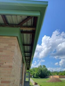 View of a brick building with a green metal roof overhang, exposed beams underneath, and a grassy field with trees and clouds in the background.