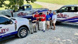 Six people stand in front of two Henley Roofing Company trucks parked on a gravel driveway, posing for a group photo outdoors.