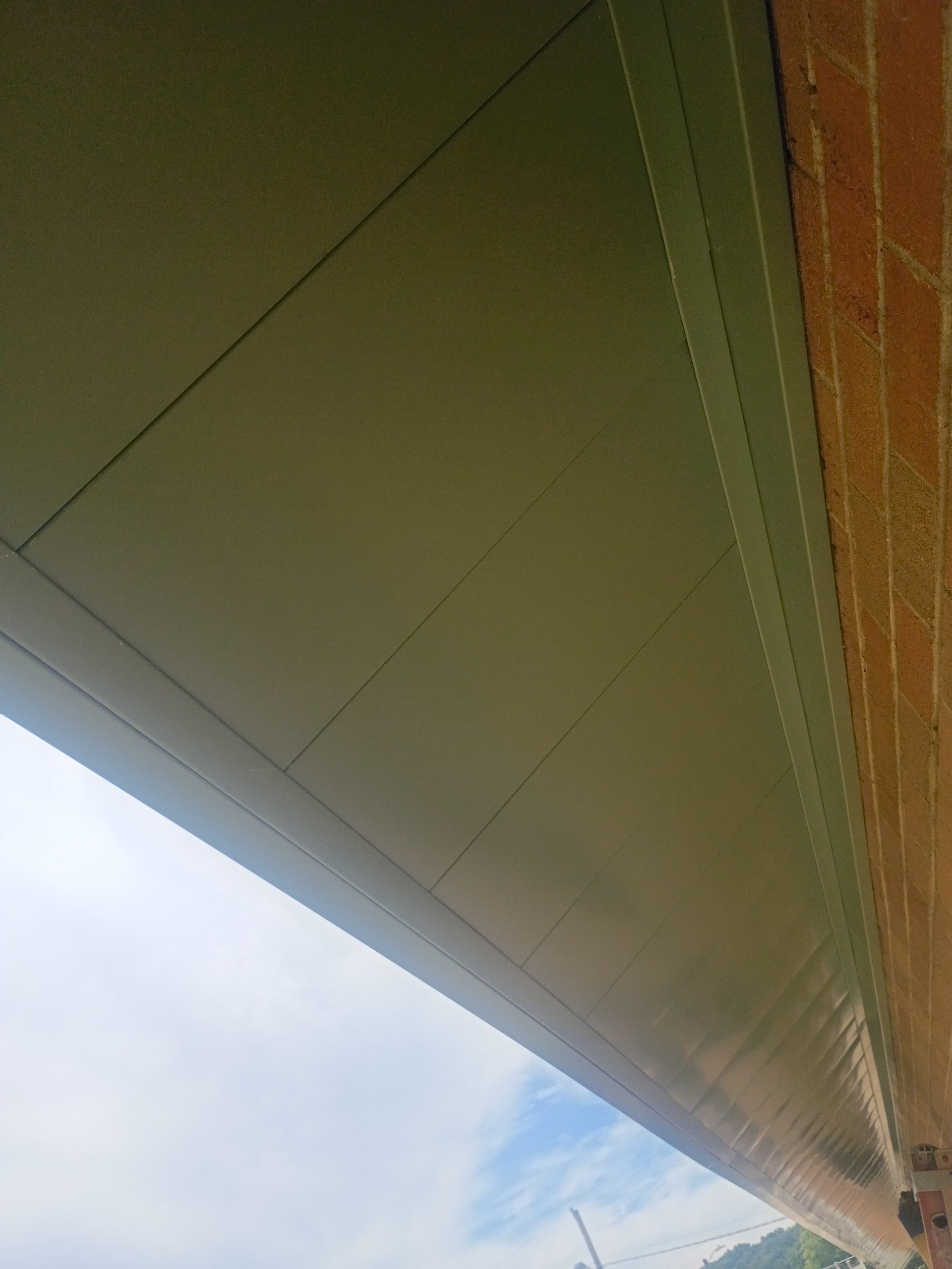 Metal roof soffit with angular panels attached to a brick wall, with a partly cloudy sky visible in the background.