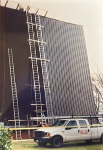 Construction workers use multiple ladders to access the side of a large corrugated metal building, with a white work truck parked nearby.