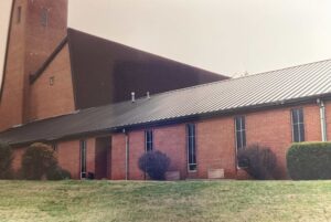 A brick church building with tall narrow windows, a metal roof, and trimmed bushes in front, seen from the side on a cloudy day.