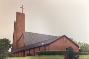 A modern red brick church with a tall, angular tower topped by a cross, surrounded by trimmed bushes and a grassy lawn under a cloudy sky.
