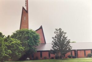 A brick church with a steep, modern angular steeple, surrounded by trees and grass under a cloudy sky.