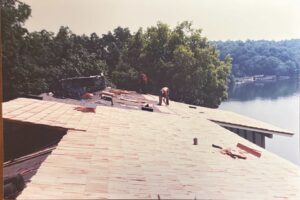 Two people work on installing roofing shingles on a house near a lake, surrounded by trees, with tools and materials scattered on the roof.