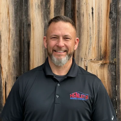 A man with a short haircut and gray beard smiles at the camera, wearing a black Henley Roofing Company polo shirt, standing in front of a wooden wall.