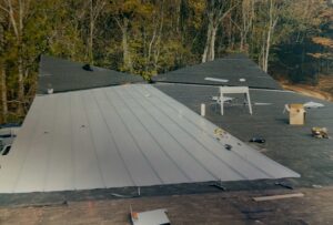 A partially completed roof shows installation in progress, with tools, materials, and metal panels laid out; trees are visible in the background.