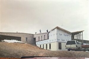 A partially constructed building with workers on the roof; walls are wrapped in Tyvek, and construction materials and a white van are nearby.
