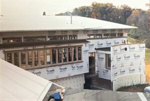 A modern house under construction with Tyvek wrap on exterior walls, large windows, and a metal roof, surrounded by trees and grass.