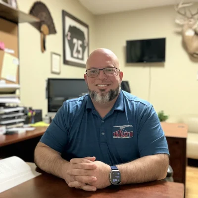 A man with glasses and a beard sits at an office desk, smiling. The office contains a computer, papers, a TV, and wall decorations including a framed jersey and a deer mount.