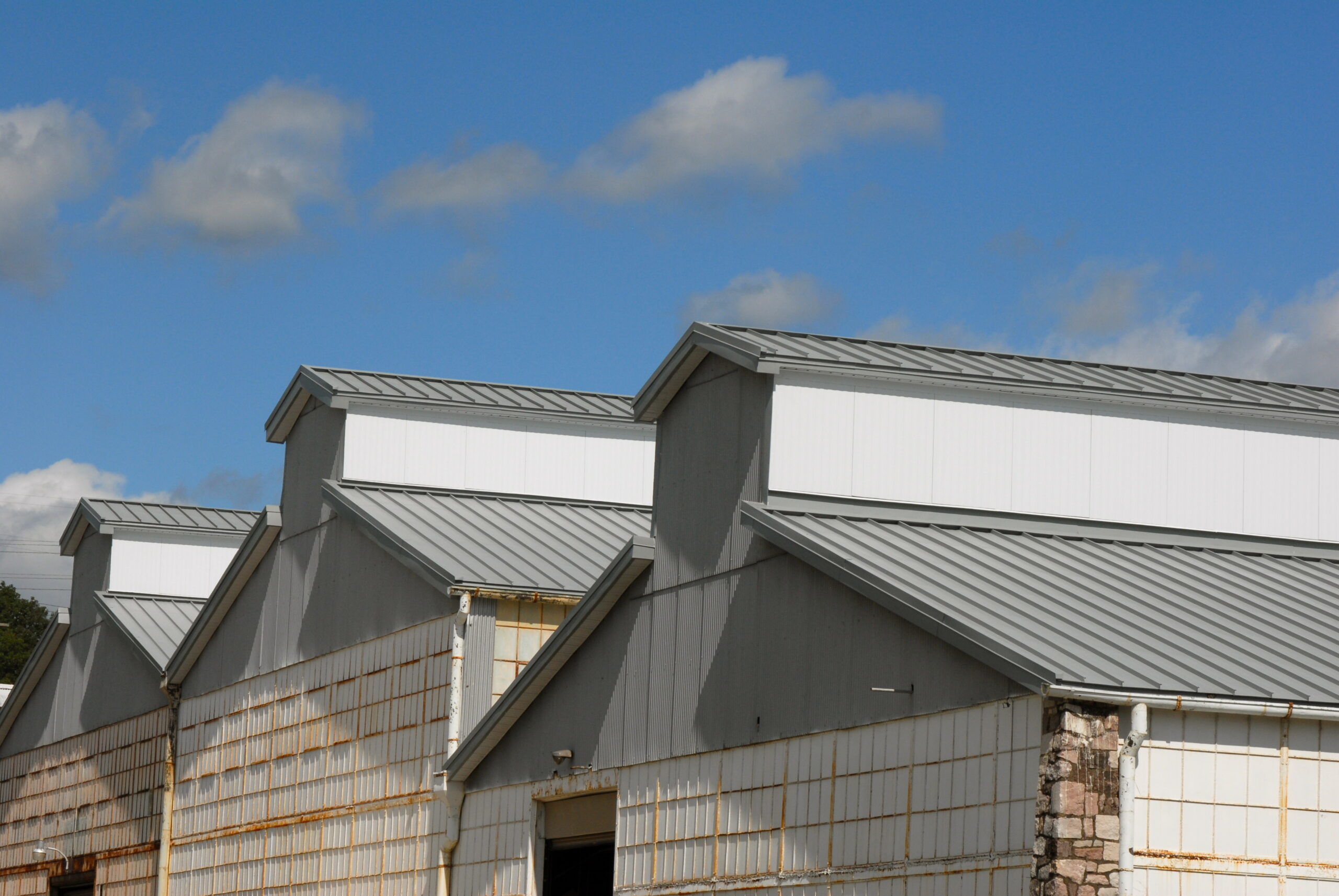 Several industrial buildings with metal roofs and white paneled walls are shown side by side under a blue sky with scattered clouds.