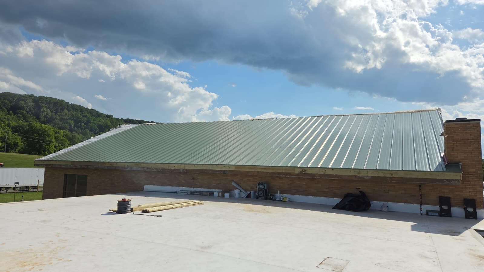 A green metal sloped roof attached to a brick building, with construction materials and tools placed on the adjacent flat white rooftop under a partly cloudy sky.