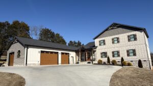 Two-story modern farmhouse with attached three-car garage, light vertical siding, brown garage doors, and a large curved driveway under a clear blue sky.