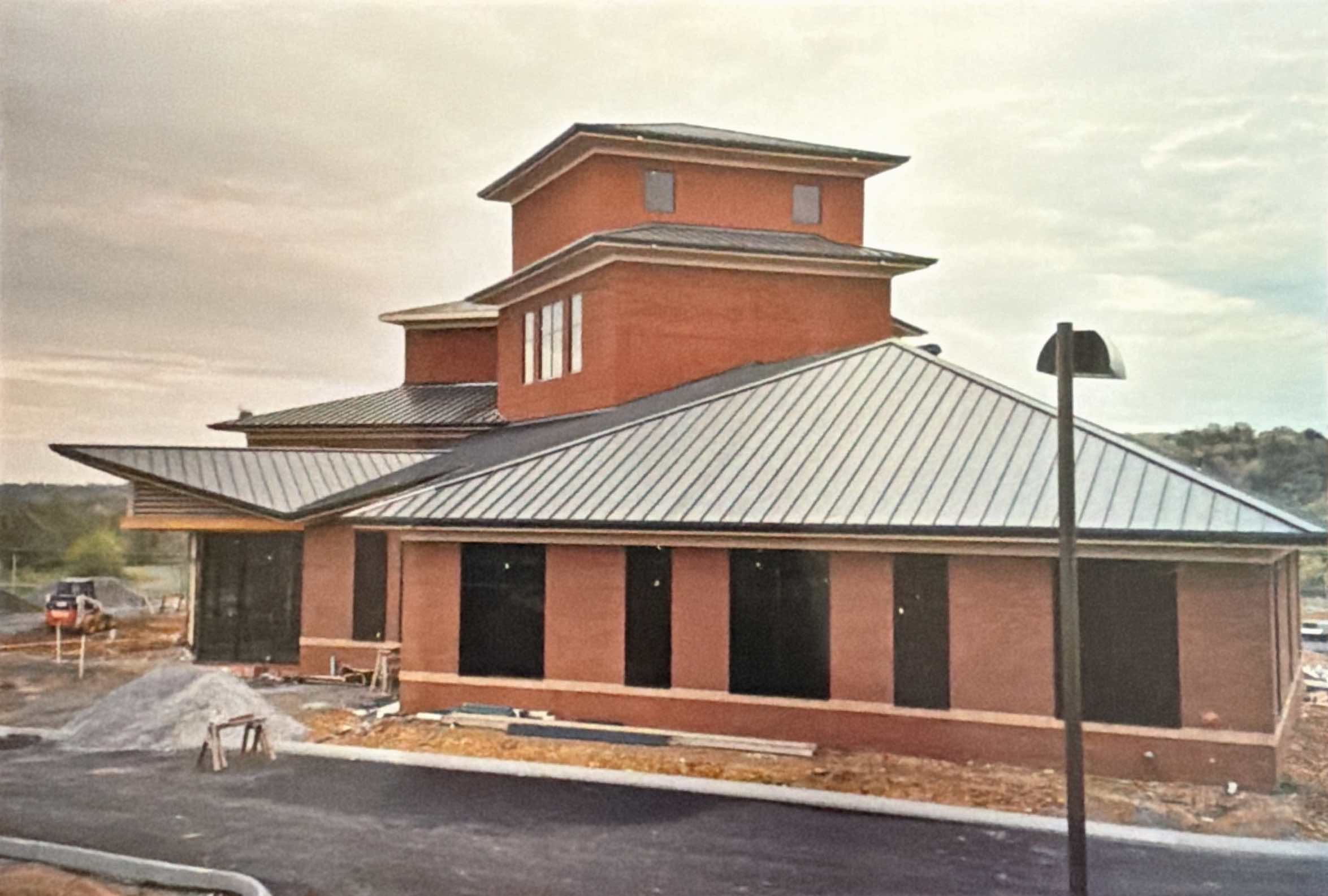 A brick building with a metal roof under construction, featuring a central tower and a surrounding paved area with construction materials visible.
