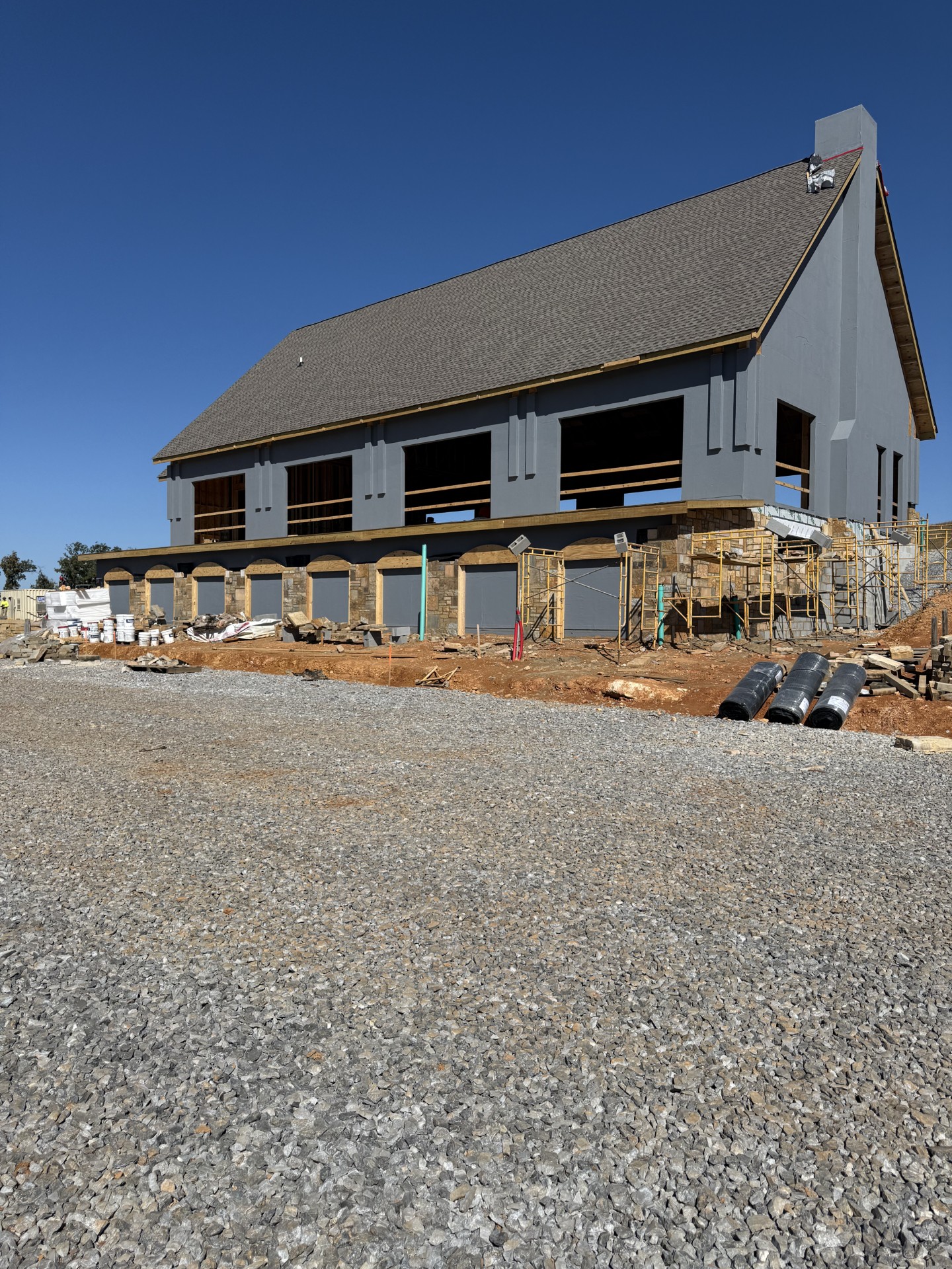 A large two-story building under construction with gray exterior walls, exposed window frames, and scaffolding, surrounded by gravel and construction materials.