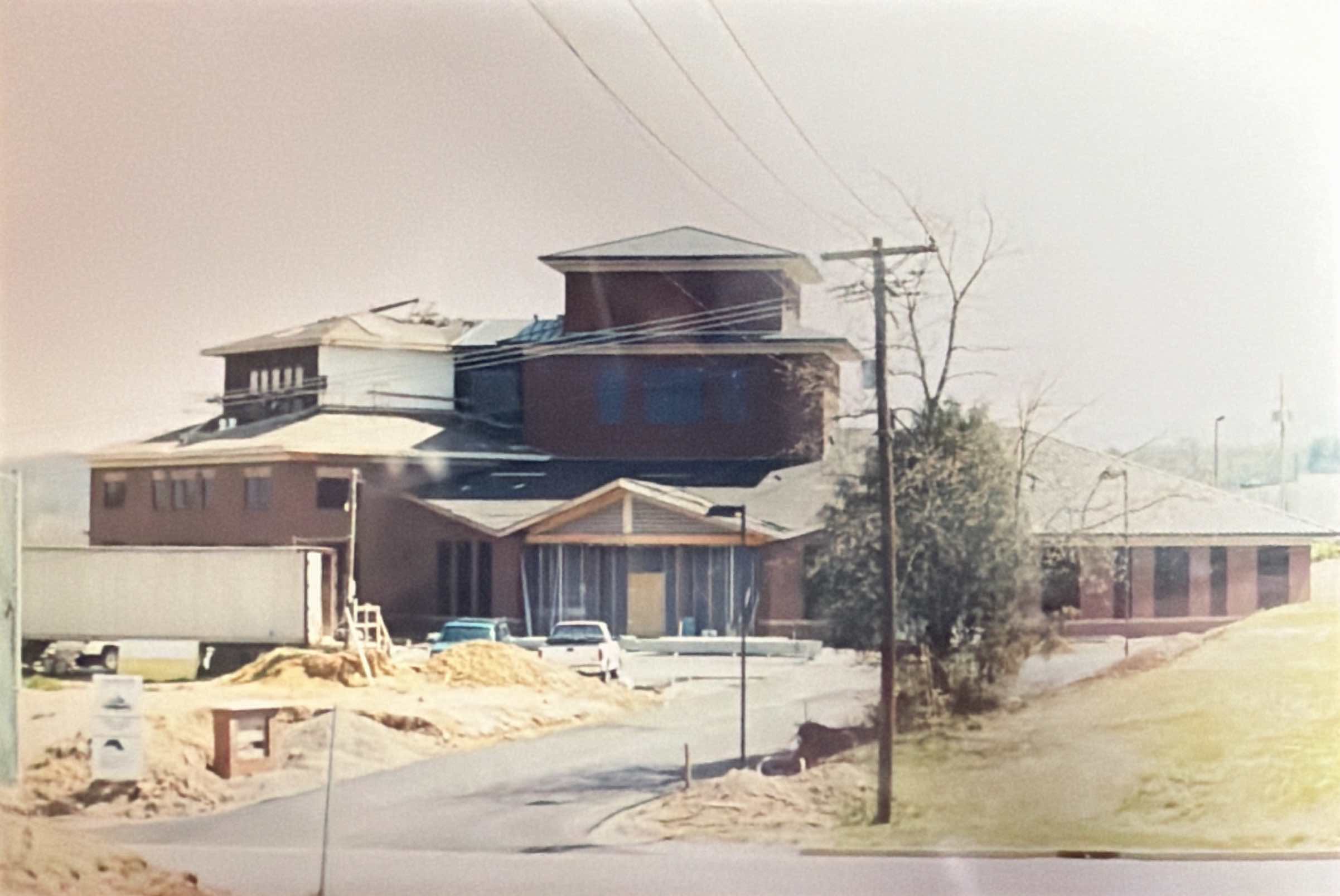 A construction site showing a large brick building with a partially finished roof, construction materials, vehicles, and a trailer nearby.