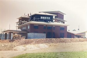 A multi-story building under construction with workers on the roof, construction equipment, and a shipping container visible on the ground.