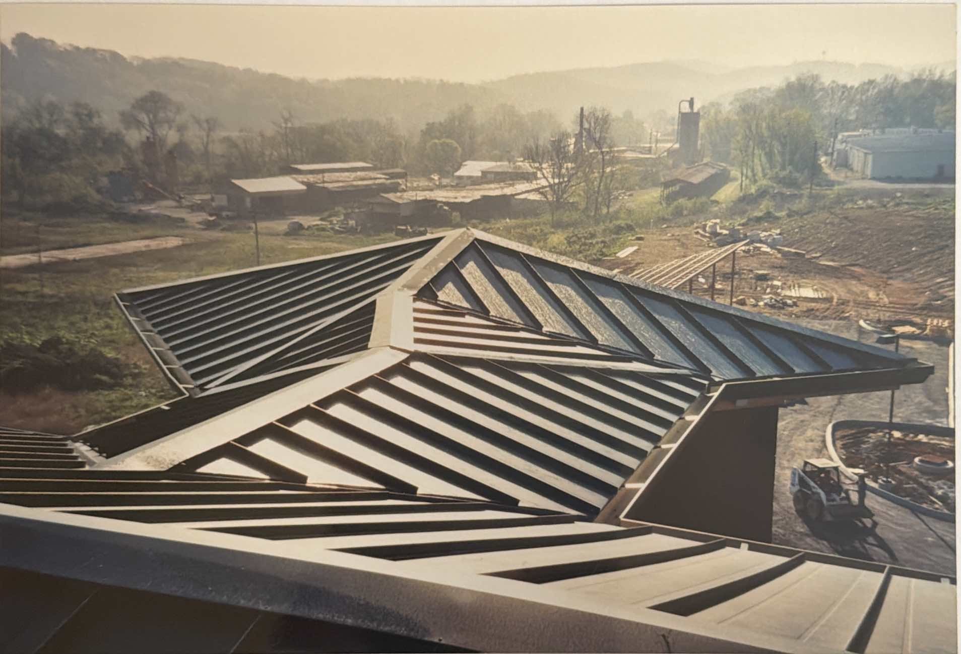 View of a modern metal roof with angular lines and ridges, overlooking a rural landscape with buildings, trees, and construction activity in the background.