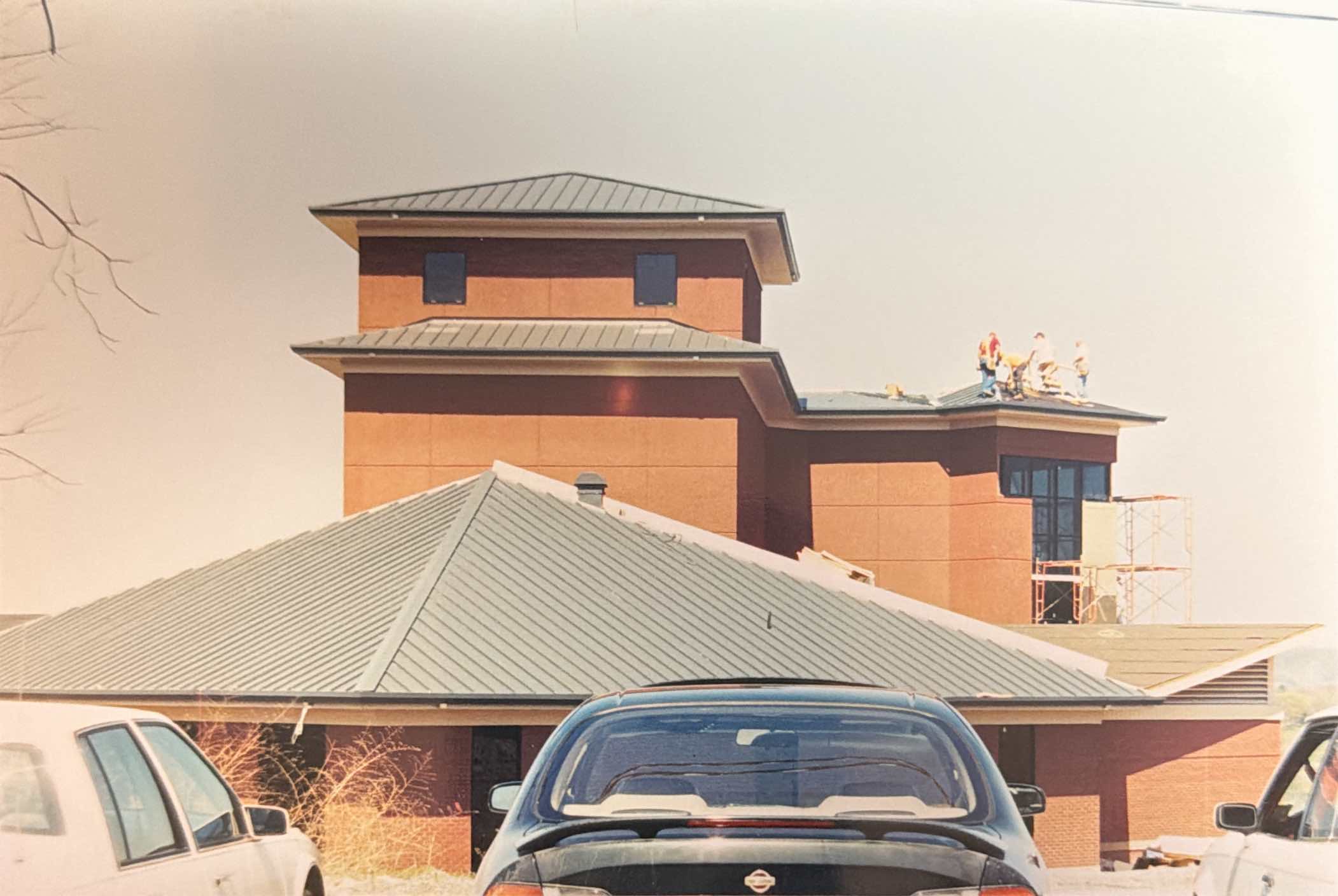 Several construction workers are working on the roof of a multi-story building, with cars parked in the foreground.