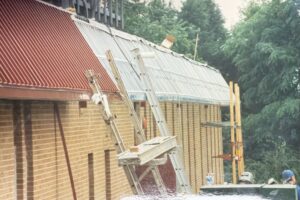 A brick building under construction with ladders and scaffolding set up against a sloped, partially metal-covered roof; trees are visible in the background.