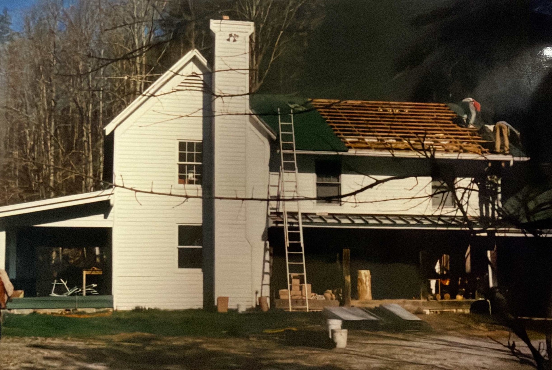A two-story white house with a carport and chimney, under roof construction; ladders are set up and a worker is visible on the exposed roof.