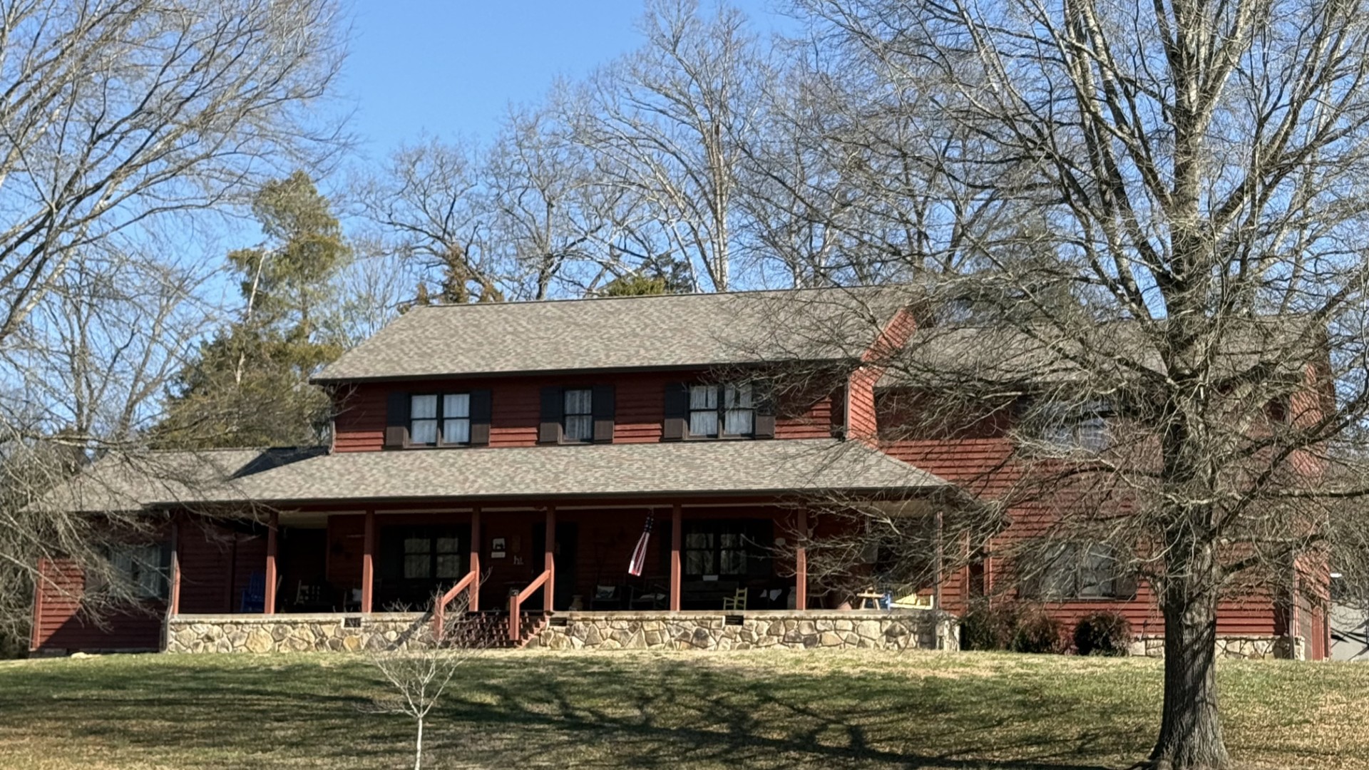 A two-story red wooden house with a stone foundation, front porch, and a sloped roof, surrounded by leafless trees and grass under a clear blue sky.