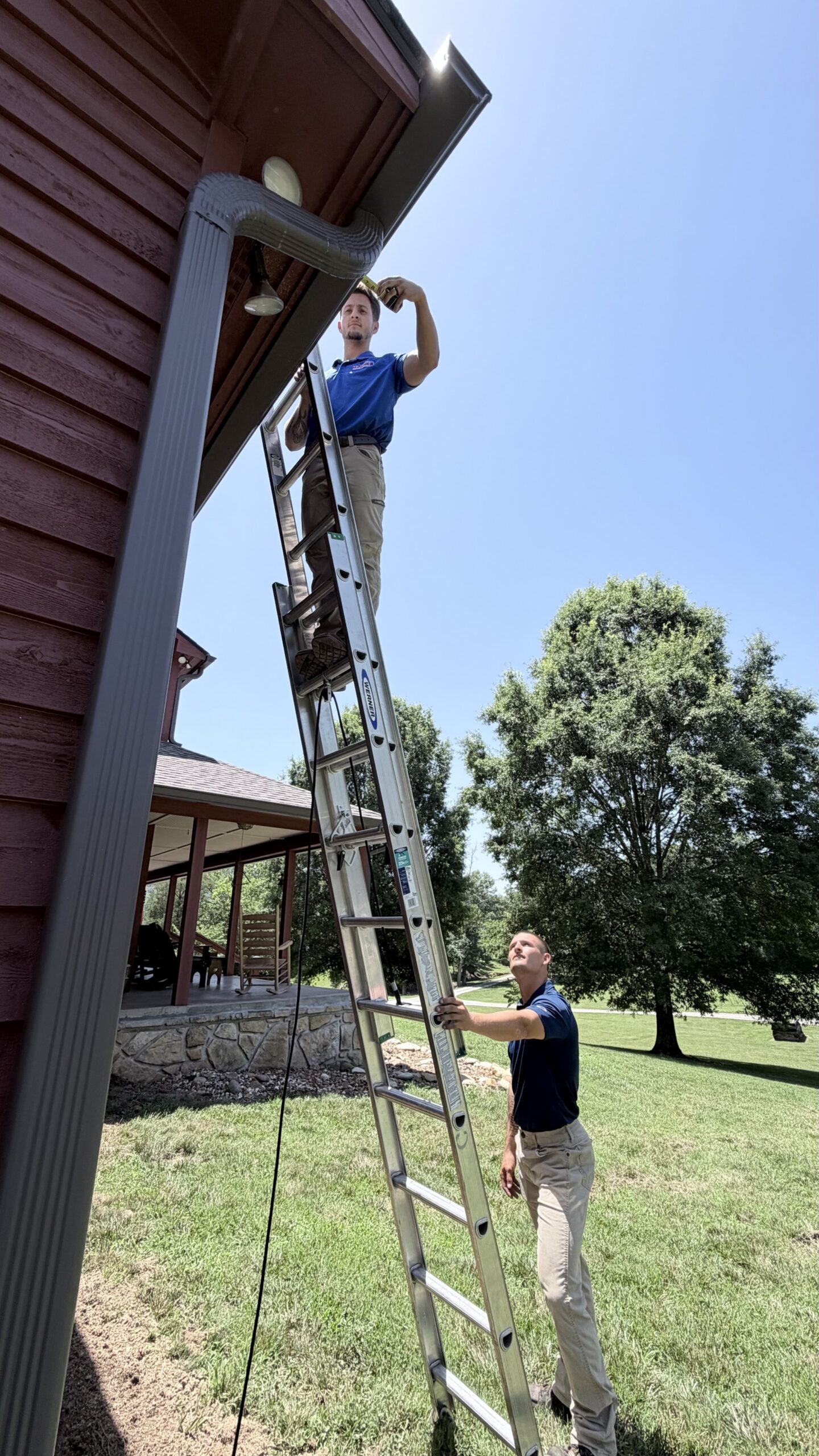 Two men work on a metal ladder by a house; one stands on the ladder inspecting the gutter while the other steadies it from below.
