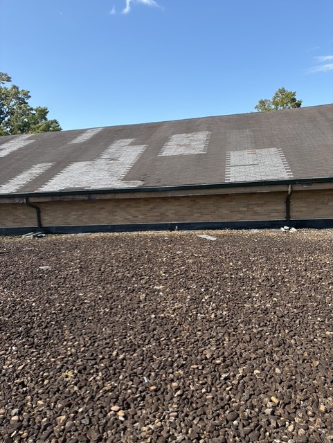 A sloped roof with visible patch repairs and missing shingles is shown behind a flat, gravel-covered surface under a clear blue sky, highlighting the importance of regular roof inspections.