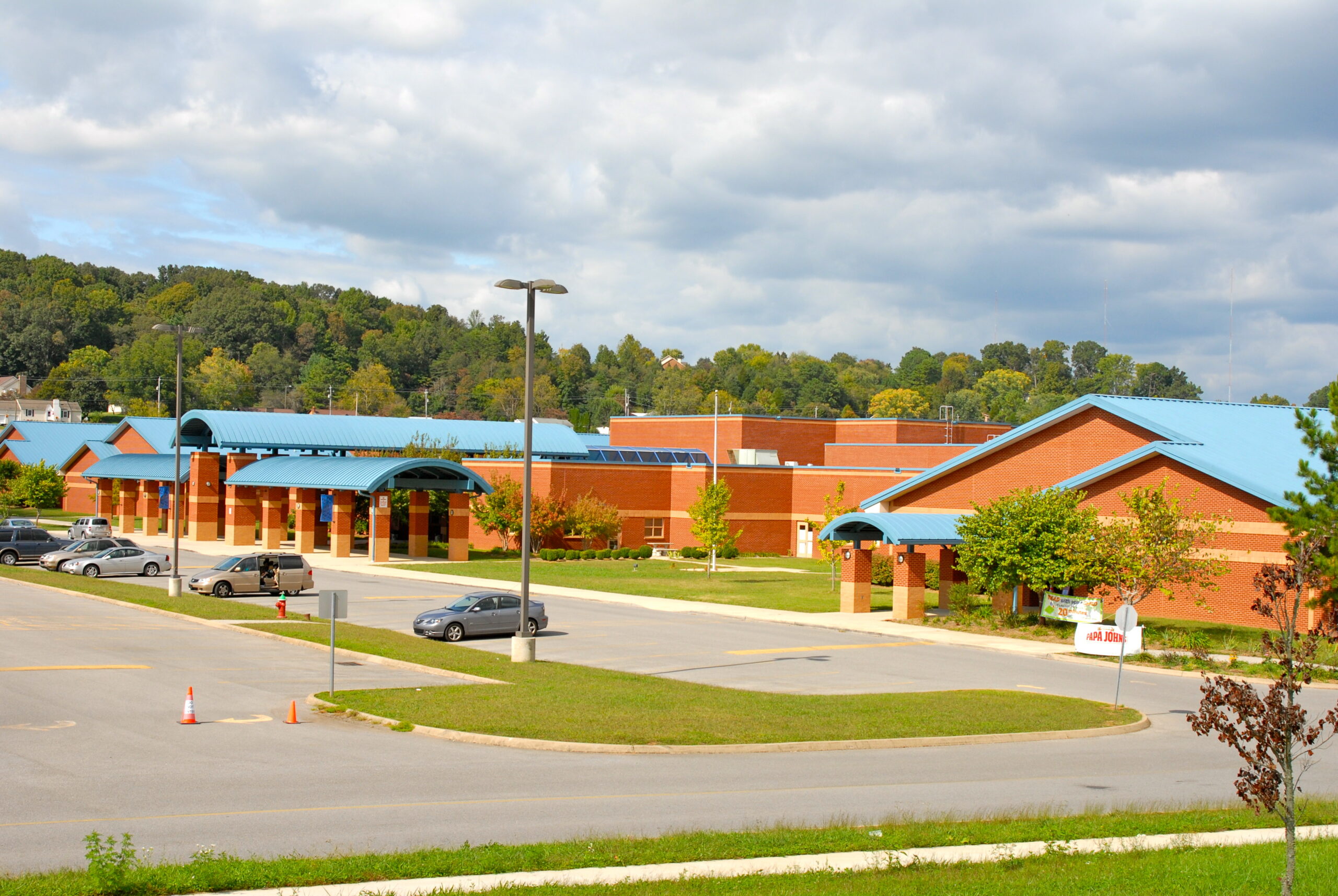 A modern brick school building with blue roofs sits beside a parking lot with several cars under a partly cloudy sky and surrounded by trees.