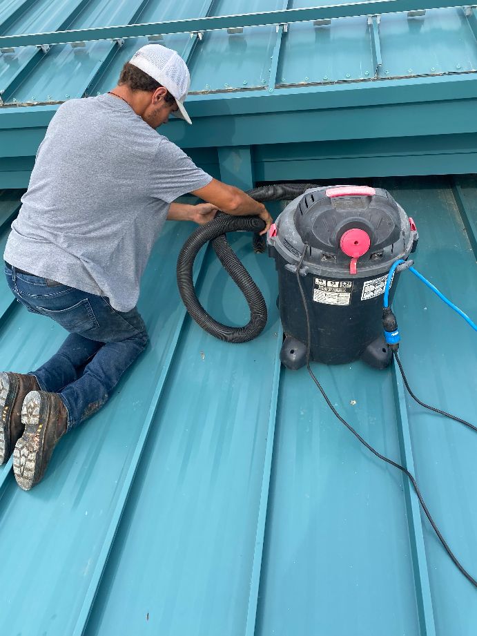 A person in a gray shirt and cap is kneeling on a blue metal roof, using a wet/dry vacuum near a vent as part of regular roof maintenance programs.