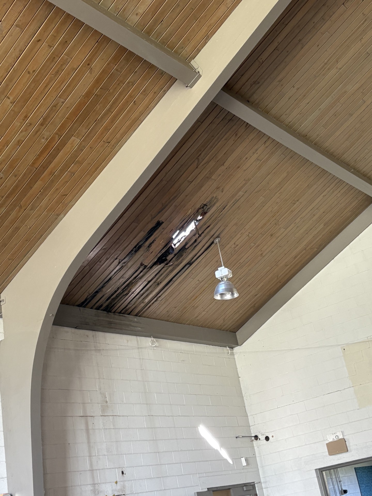 A wooden ceiling with dark water damage and staining, visible above a white brick wall in an indoor space with a hanging light fixture.