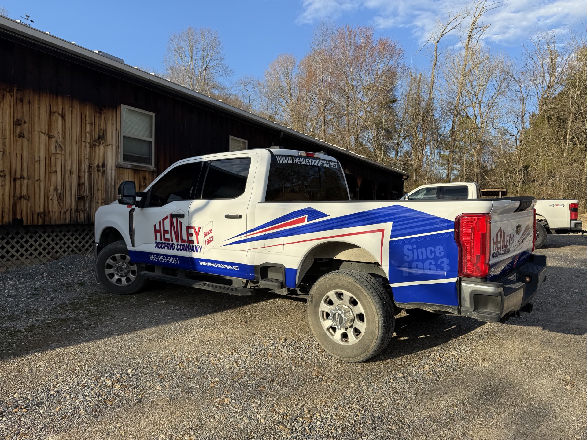 White pickup truck with Henley Roofing Company branding parked beside a wooden building on a gravel surface, with trees and a blue sky in the background.